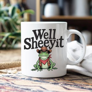 May include: White ceramic mug with the text "Well Sheeeyit" in black. The mug features a cartoon frog wearing a cowboy hat, vest, and red bandana. The frog is green with brown and red accents.
