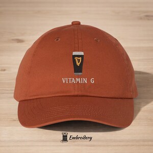 May include: A rust-colored baseball cap with the embroidered text "VITAMIN G" below a graphic of a pint of stout with a golden harp. The cap has a curved brim and a top button. The word "Embroidery" is embroidered at the bottom.