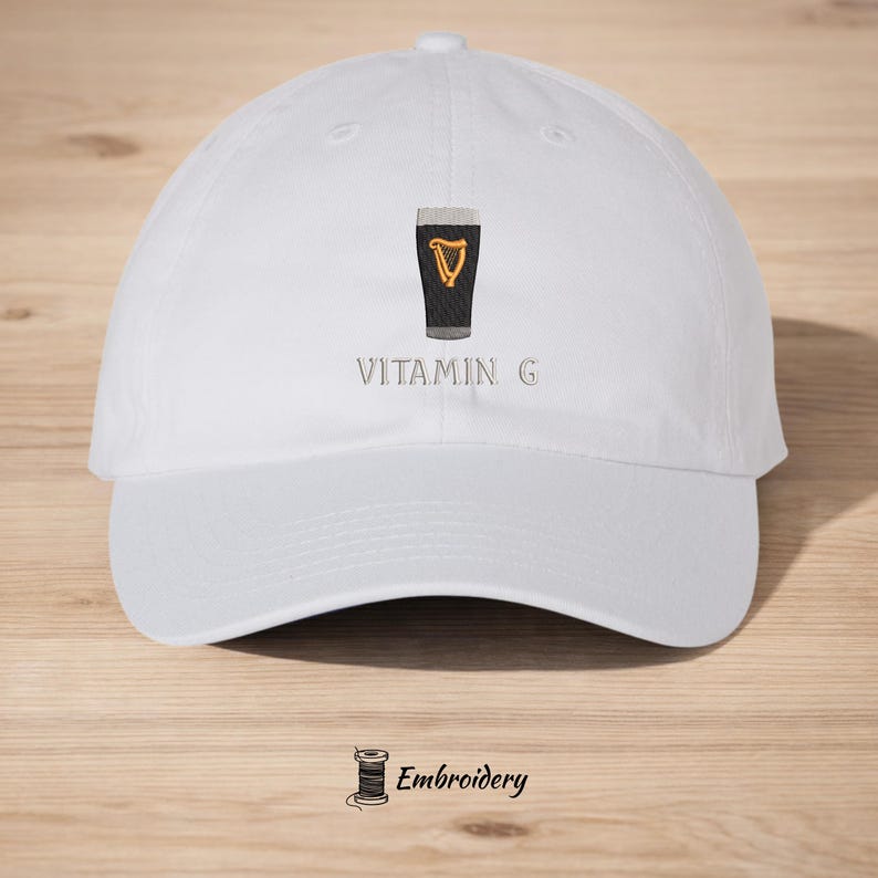 May include: White baseball cap featuring an embroidered design of a pint of stout with a golden harp and the text "Vitamin G". The cap has a curved brim and a classic six-panel construction.
