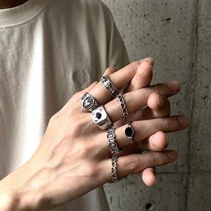 May include: A hand adorned with multiple silver rings. Designs include a ring with a playing card symbol, a ring with a black stone, and rings with various patterns and shapes. The rings are worn on multiple fingers.