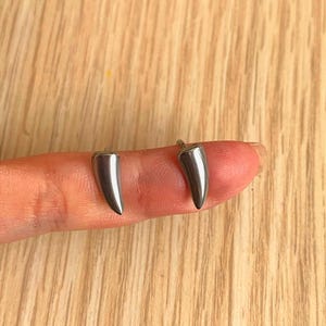 May include: Two silver-colored, tooth-shaped earrings. The earrings are small and have a polished, reflective surface. They are displayed on a finger against a light-colored wooden background. The earrings are a unique jewelry item.