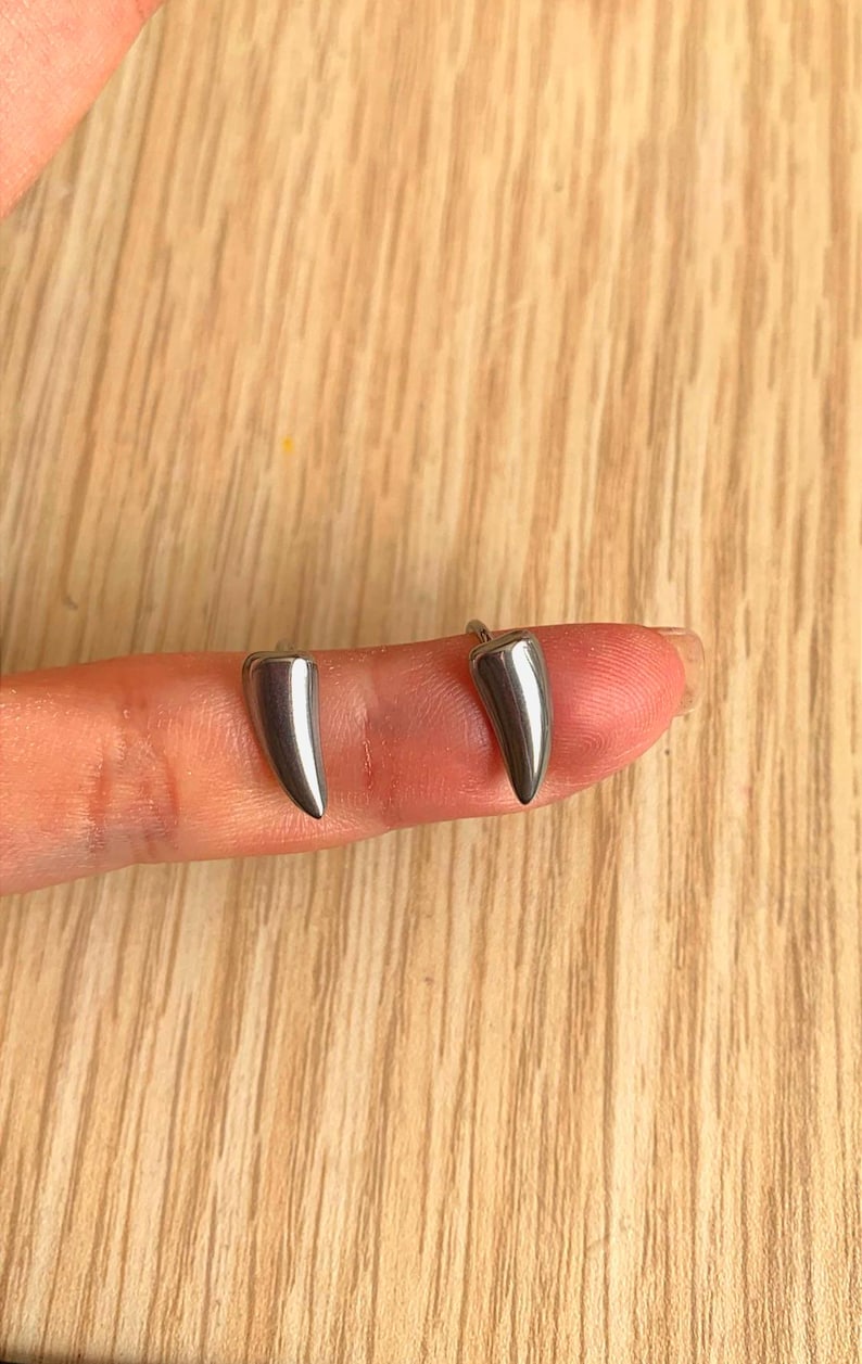 May include: Two silver-colored, tooth-shaped earrings. The earrings are small and have a polished, reflective surface. They are displayed on a finger against a light-colored wooden background. The earrings are a unique jewelry item.