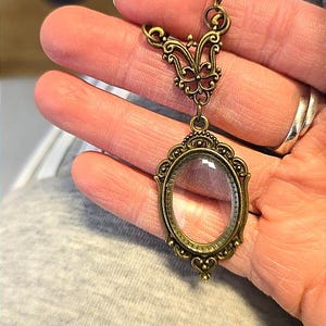 Puede incluir: Un collar con colgante de latón de estilo antiguo con un relicario ovalado de cristal. El colgante tiene detalles de filigrana ornamentados y un elemento superior decorativo. El relicario está vacío, listo para ser llenado.