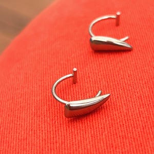 May include: Two silver-colored earrings with a curved post and a teardrop-shaped design. The earrings are set against a red background. The earrings have a polished, reflective surface.