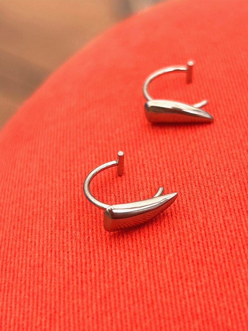 May include: Two silver-colored earrings with a curved post and a teardrop-shaped design. The earrings are set against a red background. The earrings have a polished, reflective surface.