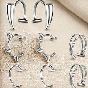 Könnte beinhalten: Eine Sammlung silberfarbener Ohrringe in verschiedenen Designs. Die Ohrringe umfassen gebogene Stile, einige mit spitzen Akzenten und andere mit einem doppelbändigen Design. Die Ohrringe werden auf einer neutralen, strukturierten Oberfläche präsentiert.