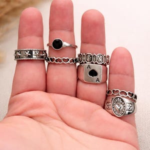 May include: A hand displays a collection of silver rings. The rings feature various designs, including stars, hearts, a black stone, and an Ace of Spades. The rings are of different sizes and styles, showcasing a variety of textures and details.