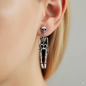 Puede incluir: Pendientes de esqueleto plateados. Los pendientes presentan un diseño de calavera y un diseño de esqueleto colgante. La caja torácica y las piernas del esqueleto son visibles. Los pendientes se llevan en la oreja de una persona.