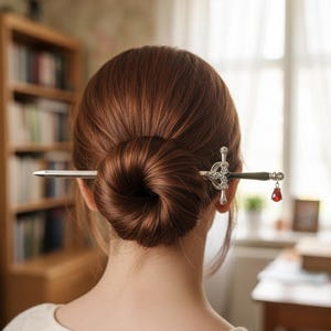 May include: A decorative hair pin featuring a sword design with a black handle and a red jewel. The hair accessory is silver and is placed in a bun of brown hair.
