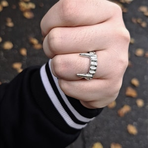 May include: A silver-colored ring with a fang design is worn on a person's finger. The ring has two prominent fangs and a series of smaller, rectangular details. The person is wearing a black jacket with white stripes on the sleeve.