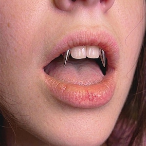 May include: Close-up of a person's mouth with silver vampire fangs. The fangs are pointed and extend from the upper teeth. The person's lips are slightly parted, revealing the tongue and teeth. The skin around the mouth is pink.