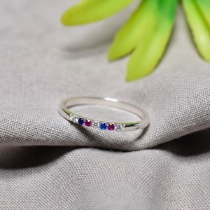 May include: A silver ring with a row of small, colorful gemstones. The ring features alternating blue, red, and clear stones. The ring is set against a gray fabric background with a green and yellow plant in the background.