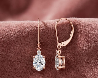 May include: Rose gold drop earrings featuring oval, clear gemstones. The earrings have a lever back closure and a small round gemstone above the oval stone. The background is a textured, pinkish-brown fabric.
