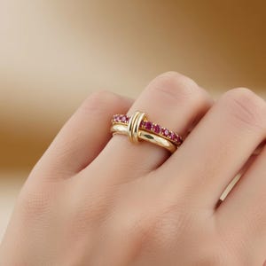 Op de afbeelding: Een paar gouden ringen, één met een rij kleine, rode edelstenen en de andere met een eenvoudige band. De ringen zijn in elkaar verweven, wat een uniek ontwerp creëert. De ringen zitten aan een vinger.
