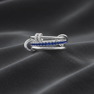 Pode incluir: Um anel de prata com um design único. O anel apresenta três bandas interligadas, uma adornada com pedras preciosas azuis e outra com pequenas gemas brancas. O anel é exibido sobre uma superfície texturizada cinza escuro.