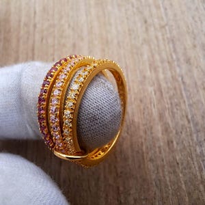 May include: A gold-colored ring with three intertwined bands. Each band is encrusted with small, colorful gemstones in shades of pink, white, and yellow. The ring is displayed on a gray surface against a wooden background.