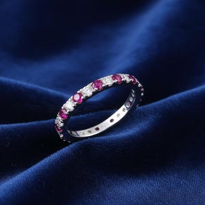 Peut inclure: Une bague d'éternité en argent ornée d'alternances de rubis ronds et de pierres précieuses claires. La bague est posée sur un fond de velours bleu foncé, mettant en valeur l'éclat des pierres. La bague est de taille 925.