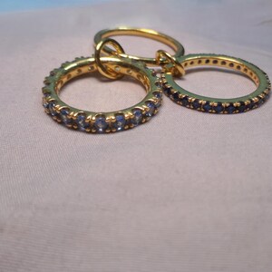 Puede incluir: Tres anillos de oro con piedras preciosas azules. Dos anillos est&aacute;n cubiertos de piedras preciosas azules, mientras que el tercero es una banda de oro lisa. Los anillos est&aacute;n entrelazados, creando un conjunto de joyas &uacute;nico.