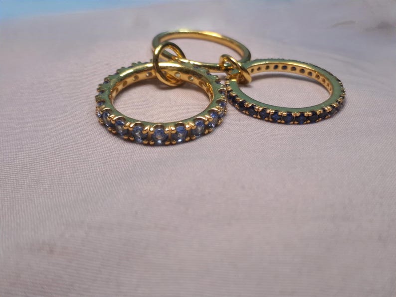 Puede incluir: Tres anillos de oro con piedras preciosas azules. Dos anillos est&aacute;n cubiertos de piedras preciosas azules, mientras que el tercero es una banda de oro lisa. Los anillos est&aacute;n entrelazados, creando un conjunto de joyas &uacute;nico.