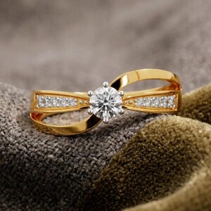 May include: A gold ring with a large, round, clear gemstone at the center. The ring has a unique design with a split band and small, clear gemstones set along the sides. The ring is displayed on a textured, gray and brown surface.