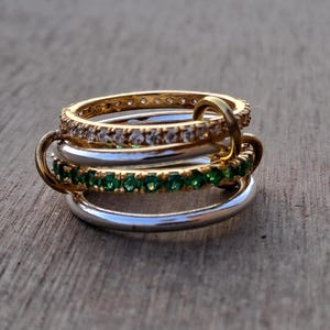 Pode incluir: Uma pilha de quatro anéis interligados. O anel superior é dourado, cravejado com pequenas pedras transparentes. Abaixo, uma faixa prateada, depois um anel dourado com pedras verdes e, finalmente, outra faixa prateada.