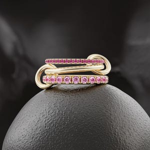 May include: Three gold rings interlocked, each adorned with a row of pink gemstones. The rings are connected by a gold clasp, creating a unique and elegant design. The jewelry is displayed on a dark, textured surface.