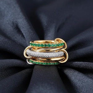 May include: A stack of gold rings with emerald green and white gemstone accents. The rings are interlocked, creating a layered effect. The gemstones are set in a pave style, adding sparkle. The rings rest on a dark blue fabric.