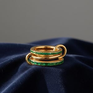 Puede incluir: Un conjunto de anillos dorados apilables con tres bandas entrelazadas. Cada banda está adornada con una fila de piedras preciosas verde esmeralda. Se exhiben sobre una tela azul oscuro.