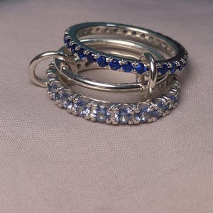Puede incluir: Tres anillos de plata entrelazados, cada uno con una banda de piedras preciosas. Un anillo presenta gemas azules, otro tiene gemas azul claro, y el tercero es una banda de plata lisa. Los anillos se exhiben sobre una tela de tono neutro.