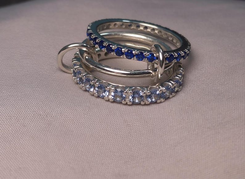 Puede incluir: Tres anillos de plata entrelazados, cada uno con una banda de piedras preciosas. Un anillo presenta gemas azules, otro tiene gemas azul claro, y el tercero es una banda de plata lisa. Los anillos se exhiben sobre una tela de tono neutro.
