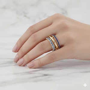 Puede incluir: Tres anillos dorados con piedras preciosas azules apilados en un dedo. Los anillos presentan una banda de peque&ntilde;as piedras azul claro, una banda dorada lisa y una banda de piedras azul oscuro. Los anillos se muestran en una mano con un fondo de m&aacute;rmol.