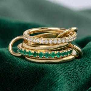 Może przedstawiać: Zestaw złotych pierścionków, z których niektóre zdobione są przezroczystymi kamieniami szlachetnymi, a inne szmaragdowozielonymi kamieniami. Pierścionki są ze sobą połączone, tworząc warstwowy efekt. Tło jest ciemnozielone.