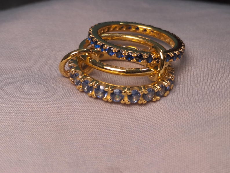 Puede incluir: Tres anillos de oro con piedras preciosas azules. Los anillos est&aacute;n apilados, con las piedras engarzadas alrededor de las bandas. Los anillos est&aacute;n sobre una superficie de color claro.