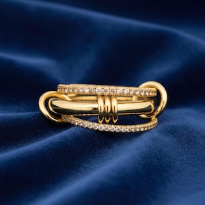 Puede incluir: Un anillo dorado con múltiples bandas, algunas incrustadas con pequeñas gemas transparentes. El anillo tiene un diseño único con elementos entrelazados, sobre un fondo azul oscuro texturizado.