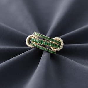 Könnte beinhalten: Ein goldener Ring mit drei ineinandergreifenden Bändern. Jedes Band ist mit kleinen, leuchtend grünen Edelsteinen besetzt. Der Ring hat ein einzigartiges Design, mit einem Abschnitt aus kleinen, klaren Edelsteinen.
