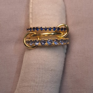 Puede incluir: Dos anillos de oro entrelazados, cada uno adornado con peque&ntilde;as piedras preciosas azules. Los anillos est&aacute;n conectados por un lazo dorado. Los anillos se muestran sobre una superficie blanca.