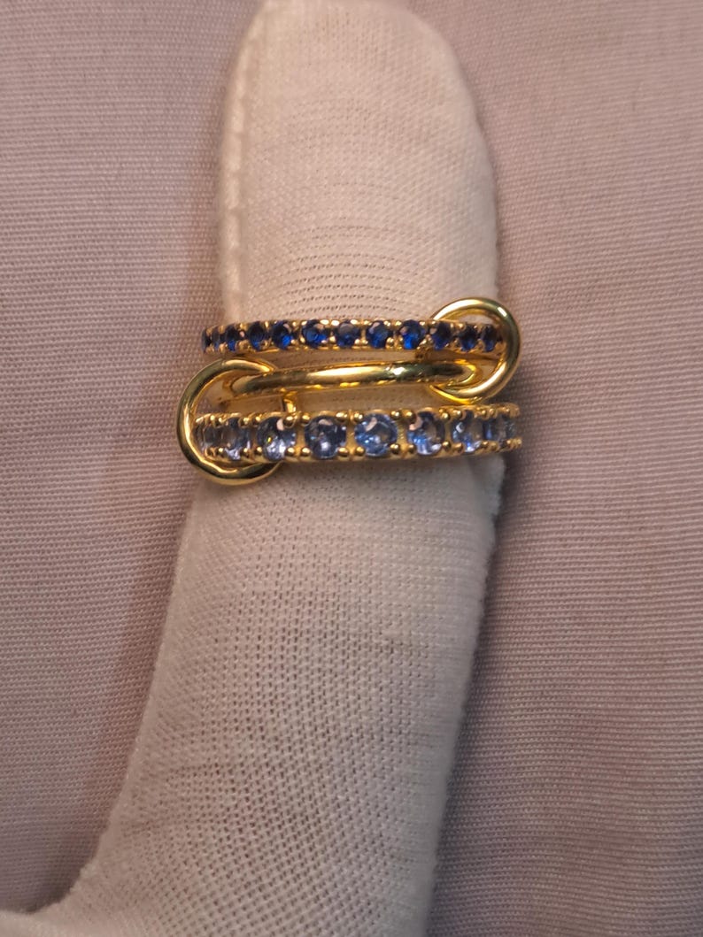 Puede incluir: Dos anillos de oro entrelazados, cada uno adornado con peque&ntilde;as piedras preciosas azules. Los anillos est&aacute;n conectados por un lazo dorado. Los anillos se muestran sobre una superficie blanca.