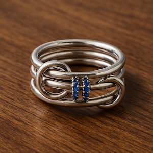 May include: A silver ring with an unusual design. The ring has multiple intertwined bands and two vertical rows of small, blue gemstones. The ring is set against a wooden surface, highlighting its metallic sheen and gemstone accents.
