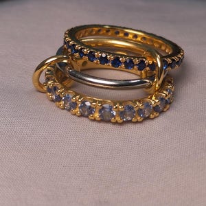 Puede incluir: Tres anillos entrelazados. Dos son dorados, uno con piedras azules, el otro con piedras claras. El tercer anillo es plateado. Los anillos se muestran sobre una superficie de color neutro.