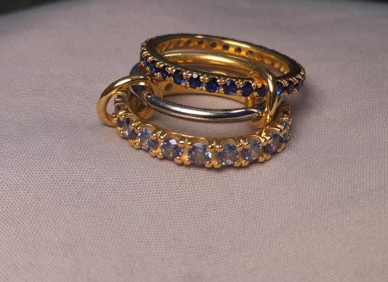 Puede incluir: Tres anillos entrelazados. Dos son dorados, uno con piedras azules, el otro con piedras claras. El tercer anillo es plateado. Los anillos se muestran sobre una superficie de color neutro.
