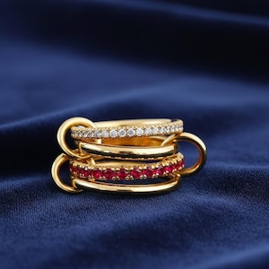 May include: A stack of gold rings with a connecting loop design. One ring is encrusted with small white gemstones, another with red gemstones, and a third is plain gold. The rings are displayed on a dark blue fabric.