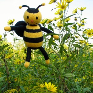 May include: A knitted bee toy with a black and yellow striped body, black wings, and a smiling face. The bee is positioned amongst tall green plants and bright yellow flowers, creating a whimsical scene.