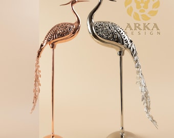 Puede incluir: Dos esculturas decorativas de pájaros de metal. Una es de color cobre, la otra plateada. Ambas tienen cuellos largos, cuerpos detallados y largas plumas de cola fluidas. Se alzan sobre postes delgados con bases redondas. El logotipo "ARKA DESIGN" es visible.