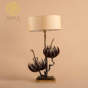 Peut inclure: Une lampe de table d&eacute;corative avec un abat-jour beige et une base &agrave; motif floral de couleur bronze. La base pr&eacute;sente des formes stylis&eacute;es en forme de fleurs et des d&eacute;tails complexes. La lampe est con&ccedil;ue pour une utilisation en int&eacute;rieur.