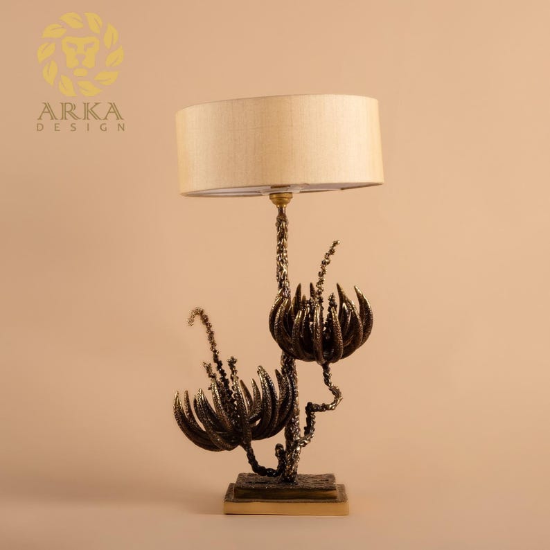 Peut inclure: Une lampe de table d&eacute;corative avec un abat-jour beige et une base &agrave; motif floral de couleur bronze. La base pr&eacute;sente des formes stylis&eacute;es en forme de fleurs et des d&eacute;tails complexes. La lampe est con&ccedil;ue pour une utilisation en int&eacute;rieur.