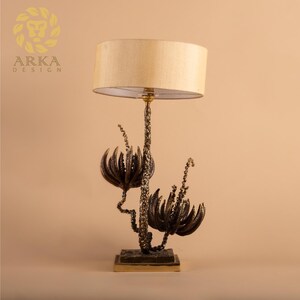 Peut inclure: Lampe de table d&eacute;corative avec abat-jour beige et base florale de couleur bronze. La base pr&eacute;sente des motifs stylis&eacute;s de fleurs et de tiges. La lampe est sur une base carr&eacute;e et a un accent dor&eacute;.