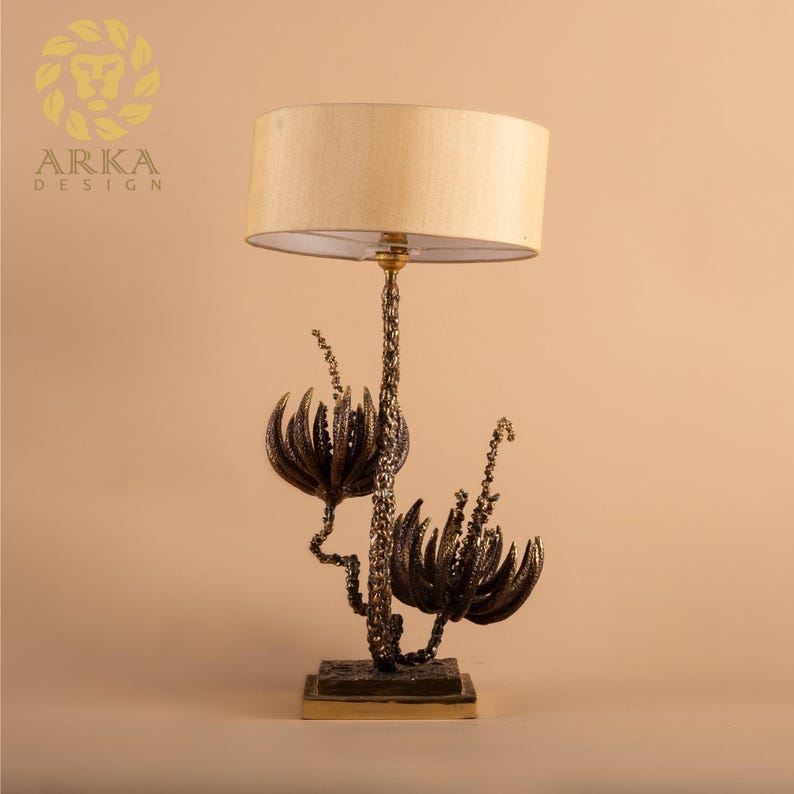 Peut inclure: Lampe de table d&eacute;corative avec abat-jour beige et base florale de couleur bronze. La base pr&eacute;sente des motifs stylis&eacute;s de fleurs et de tiges. La lampe est sur une base carr&eacute;e et a un accent dor&eacute;.