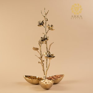 Könnte beinhalten: Ein goldfarbenes dekoratives Centerpiece mit floralem Design. Das Centerpiece hat drei kleine Schalen an der Basis, gefüllt mit Nüssen. Das florale Design hat einen astähnlichen Stiel mit Blüten und Blättern. Das Logo "ARKA DESIGN" befindet sich in der oberen rechten Ecke.