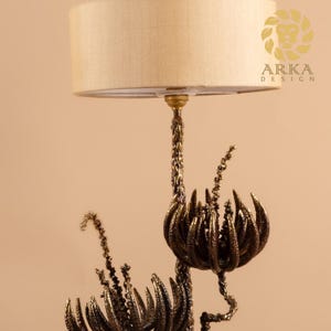 Peut inclure: Une lampe de table d&eacute;corative avec un abat-jour beige et une base de couleur bronze. La base pr&eacute;sente un design organique complexe avec des &eacute;l&eacute;ments floraux et v&eacute;g&eacute;taux. La lampe est con&ccedil;ue par Arka Design.