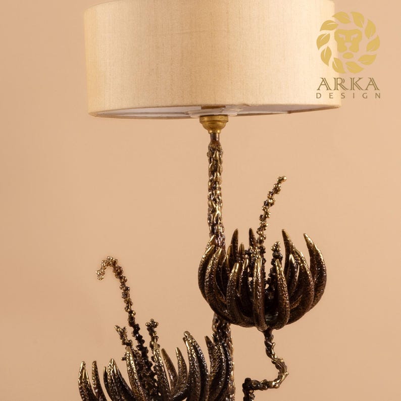 Peut inclure: Une lampe de table d&eacute;corative avec un abat-jour beige et une base de couleur bronze. La base pr&eacute;sente un design organique complexe avec des &eacute;l&eacute;ments floraux et v&eacute;g&eacute;taux. La lampe est con&ccedil;ue par Arka Design.